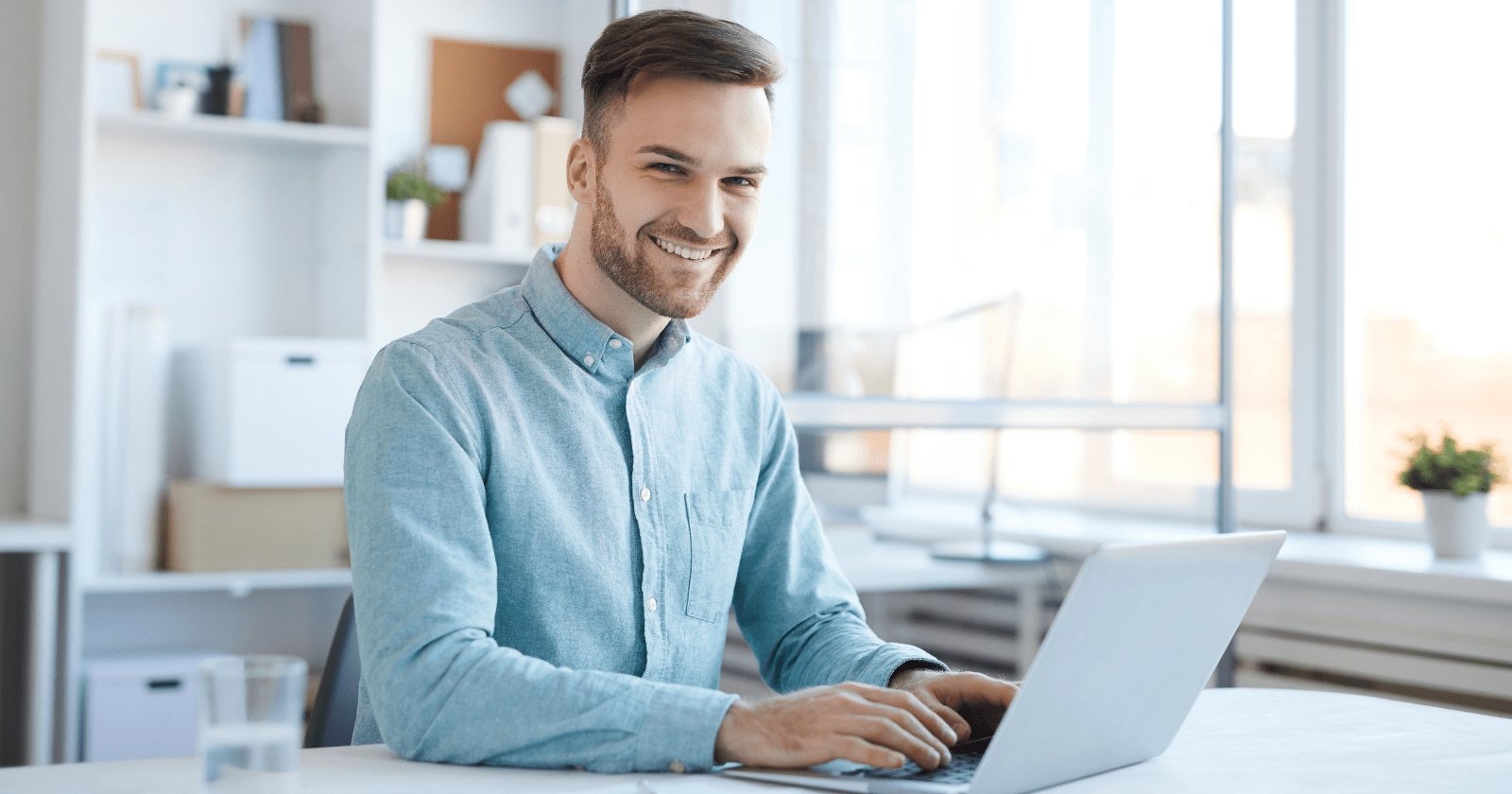 The photo shows a smiling man sitting in front of a laptop.