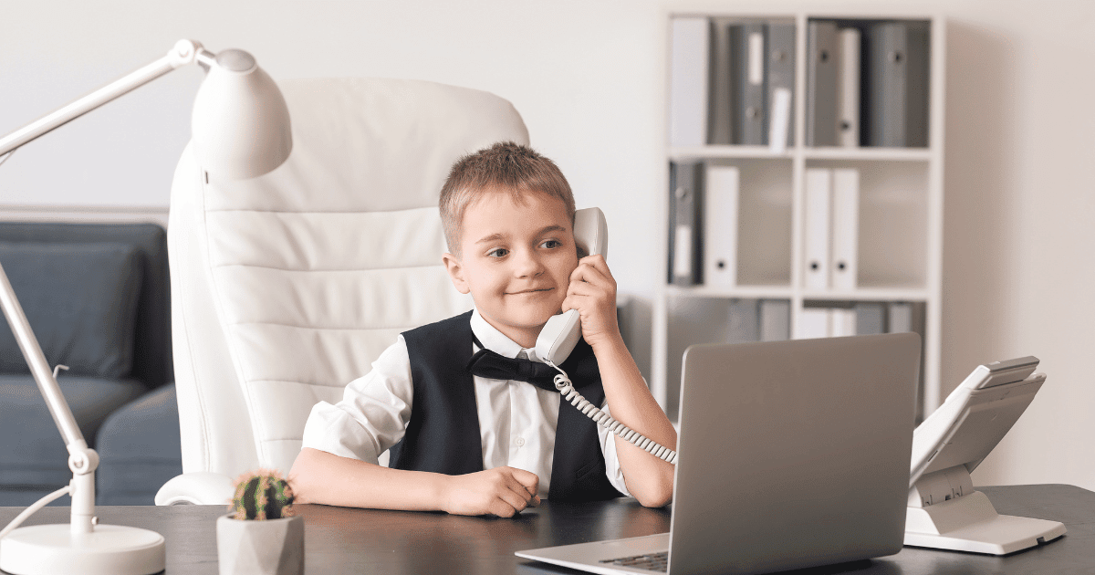 In the photo, a child in a suit sits at a desk in front of an open laptop and takes a phone call