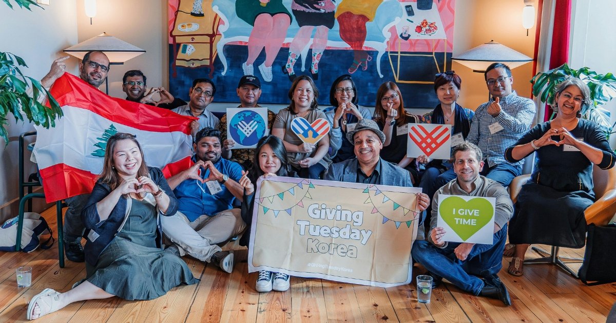 Grupp kbir ta’ voluntiera minn ħafna pajjiżi jippożaw għal ritratt. Għandhom bandiera Lebaniża, banner tal-kartun bl-islogan ‘Giving Tuesday Korea’ u kartellun żgħir b’qalb u l-kliem ‘Nagħti l-ħin’.