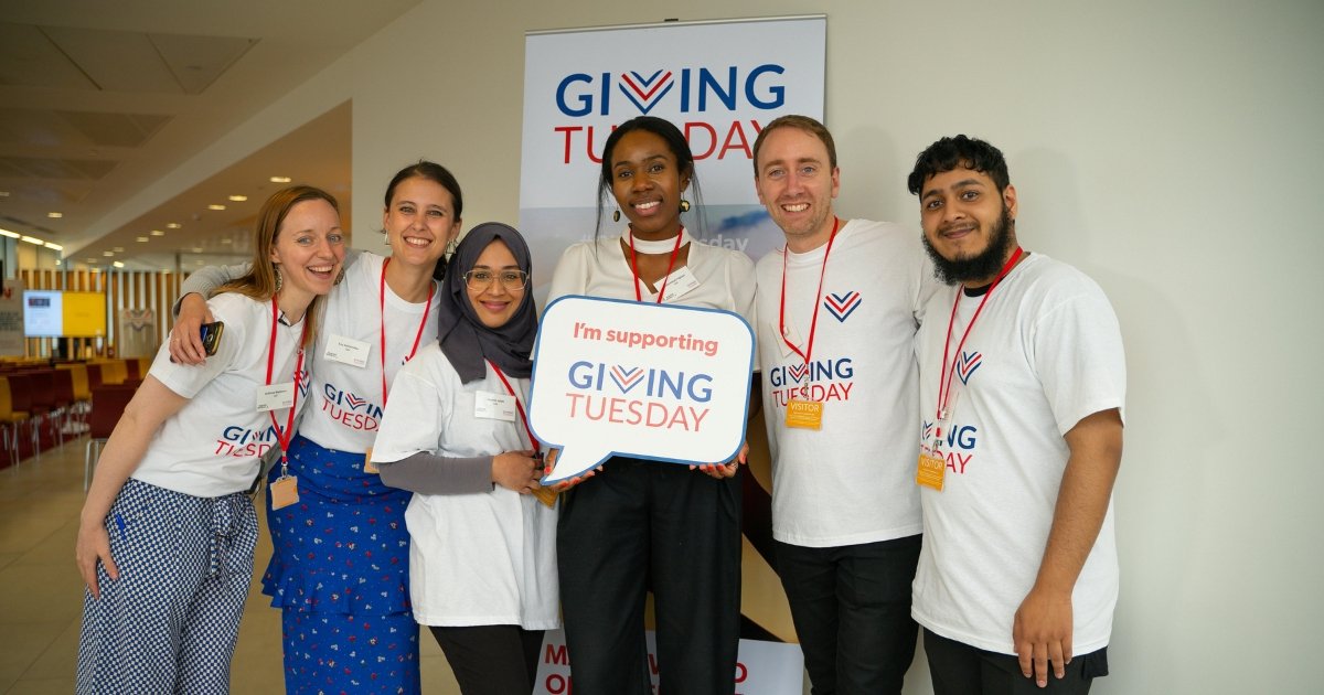 Fir-ritratt jidher grupp ta’ voluntiera ta’ nazzjonalitajiet differenti, jippożaw mal-ħajt bil-logo Giving Tuesday. Qegħdin iżommu sinjal tal-kartun bl-islogan ‘Qed nappoġġja Giving Tuesday’.